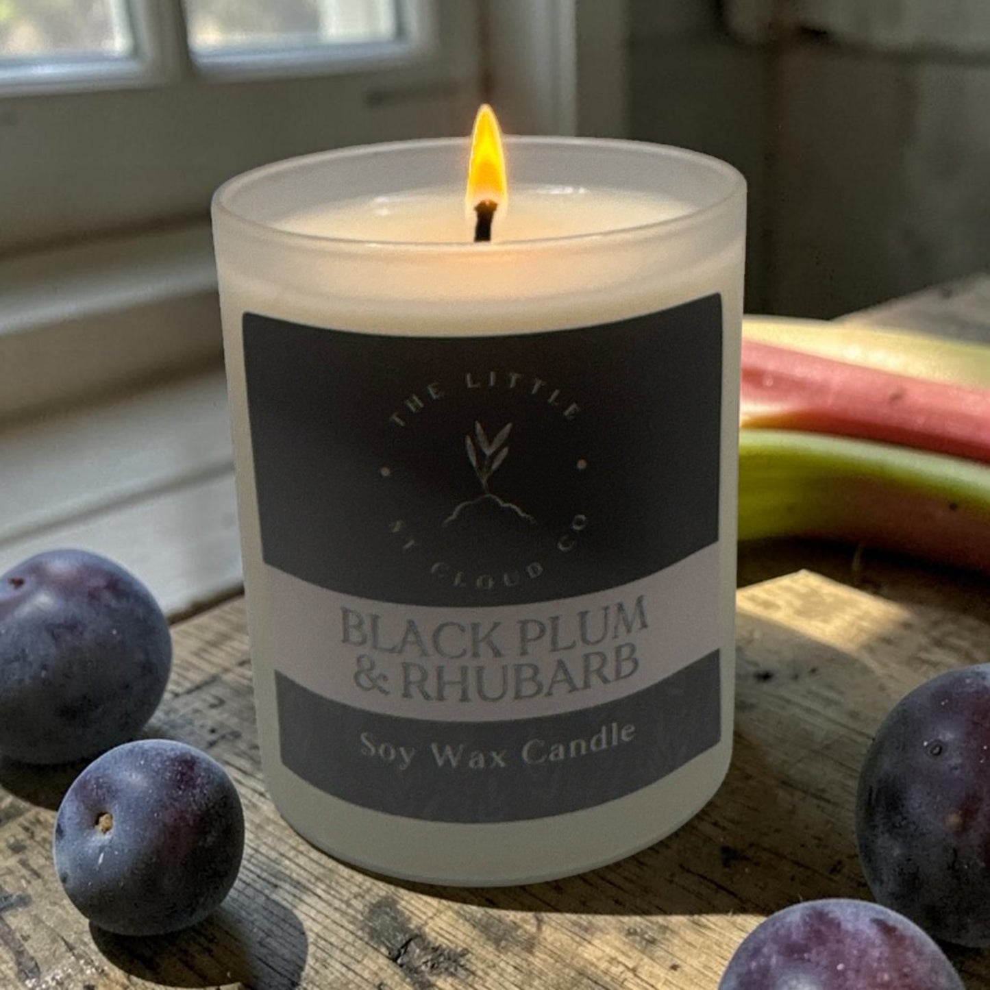 Soy wax candle labeled 'Black Plum & Rhubarb' with plums on a wooden surface.