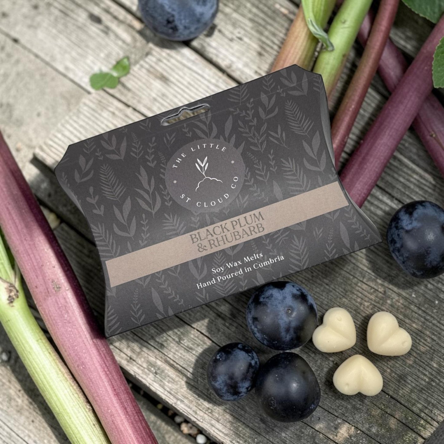 Packaging of Black Plum & Rhubarb soy wax melts on a wooden surface with plums and rhubarb.