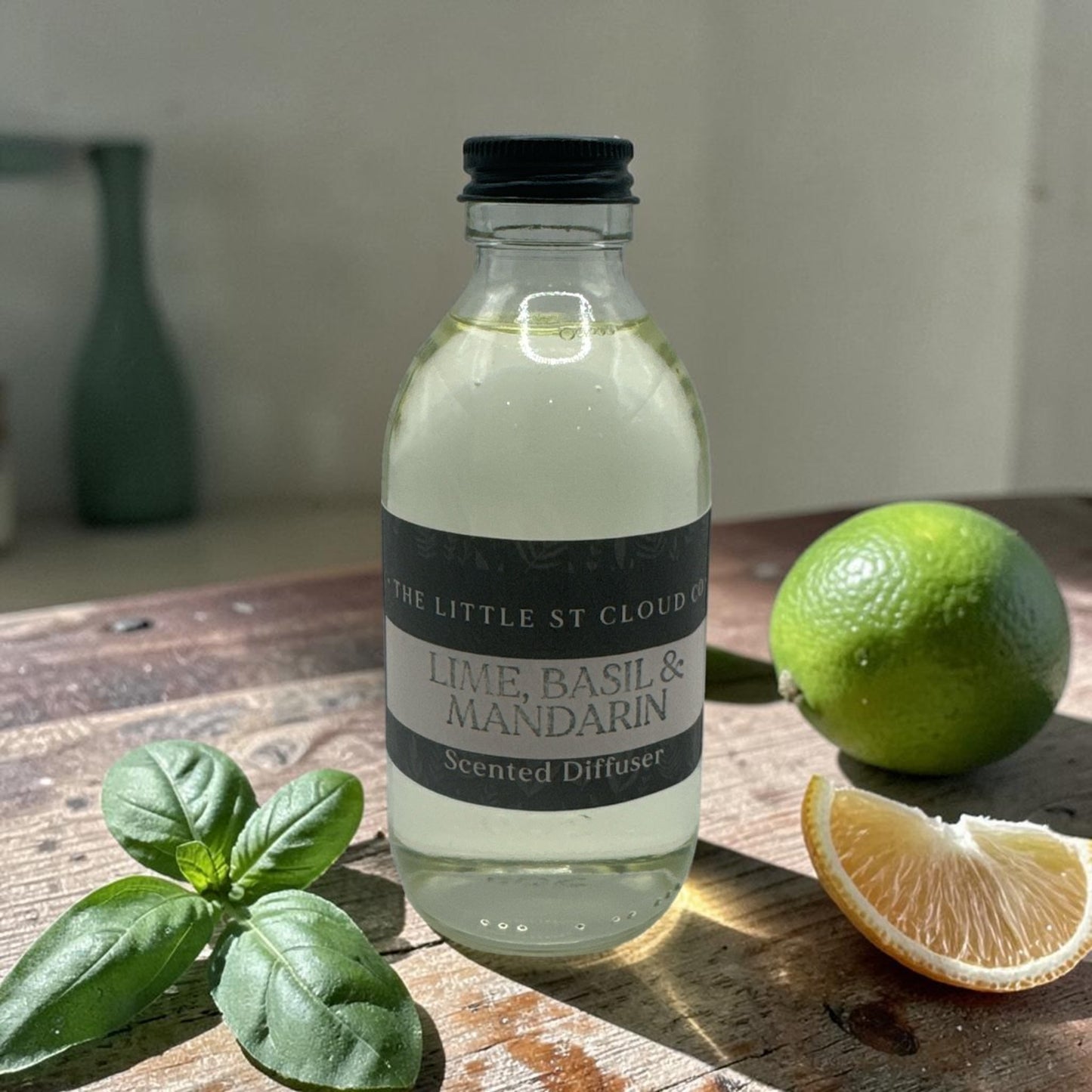 Bottle of 'Lime, Basil & Mandarin' scented diffuser on a wooden surface with a lime and basil leaves.