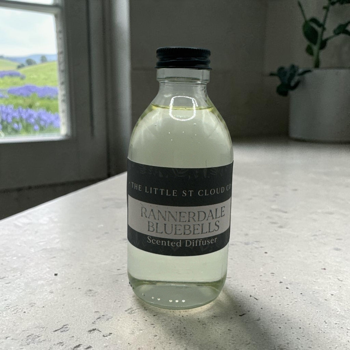A small clear glass bottle of Rannerdale Bluebells scented diffuser liquid by The Little St Cloud Co, sitting on a light speckled countertop near a window.