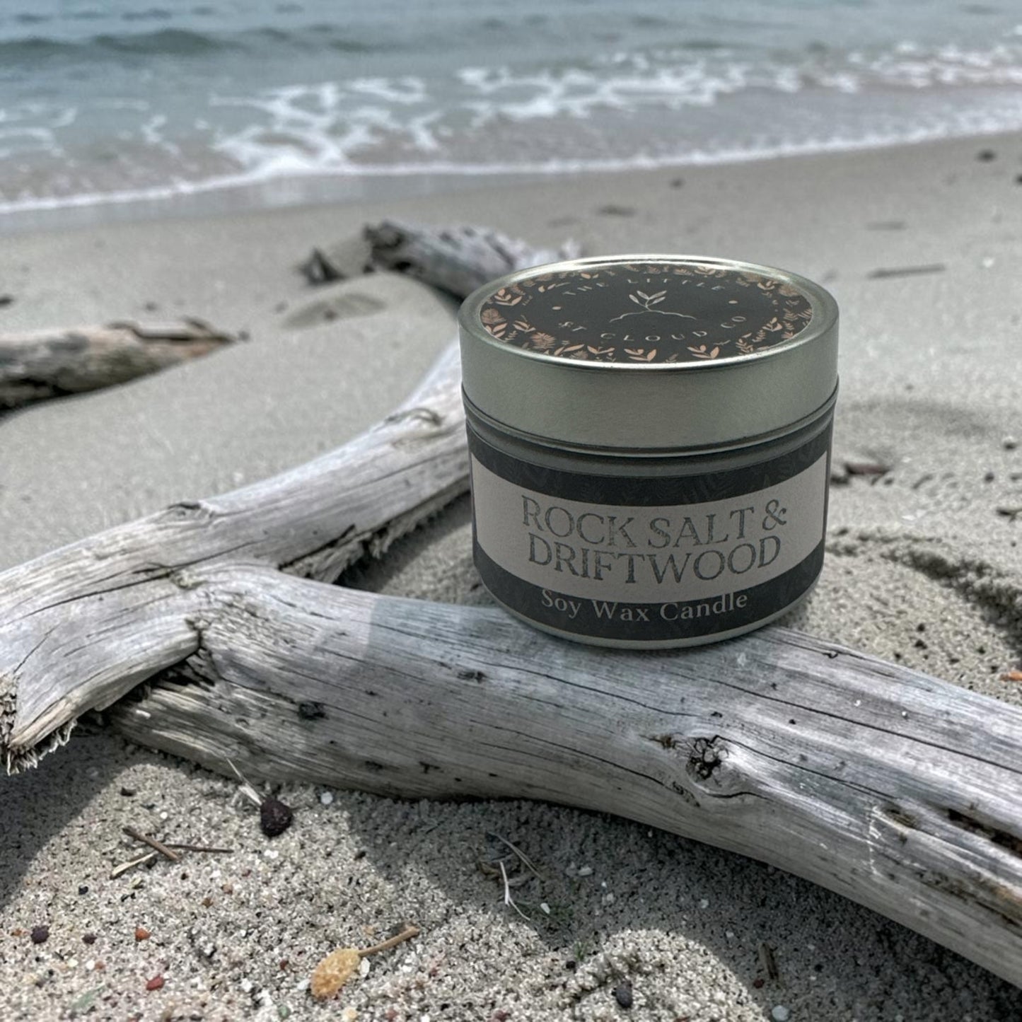 Candle jar labeled 'Rock Salt & Driftwood' on a beach with driftwood and ocean waves in the background.