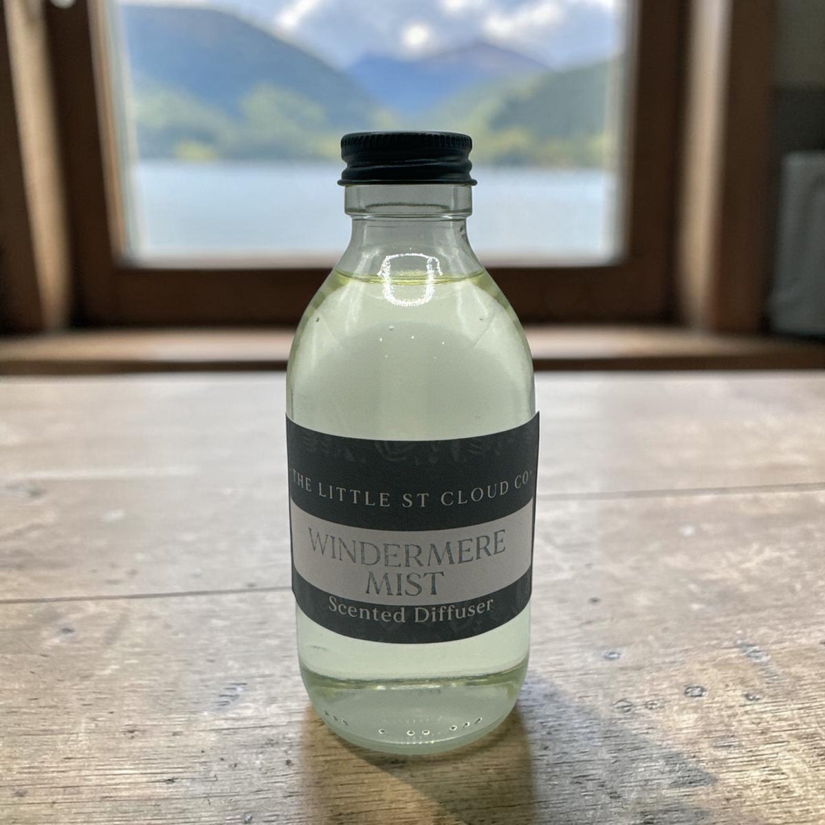 a clear glass bottle of diffuser refill by the little st cloud co in a window with lake views