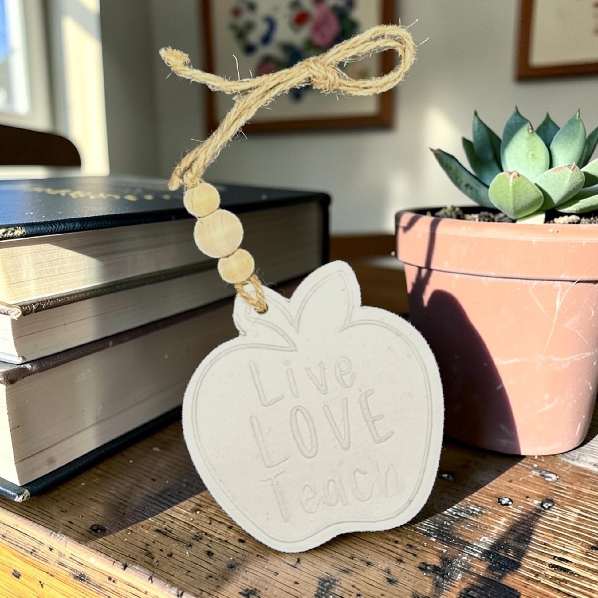 Decorative apple-shaped tag with 'Live Love Teach' text on a wooden surface with books and a plant in the background.