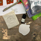 Small bag, bottle, and decorative item on a wooden surface with books in the background
