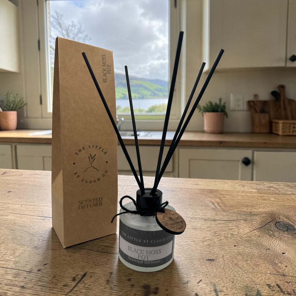 A reed diffuser bottle with black reeds and a wooden tag sits on a wooden table next to its brown kraft paper packaging box. The background shows a kitchen environment with a window looking out onto a lake and mountains.
