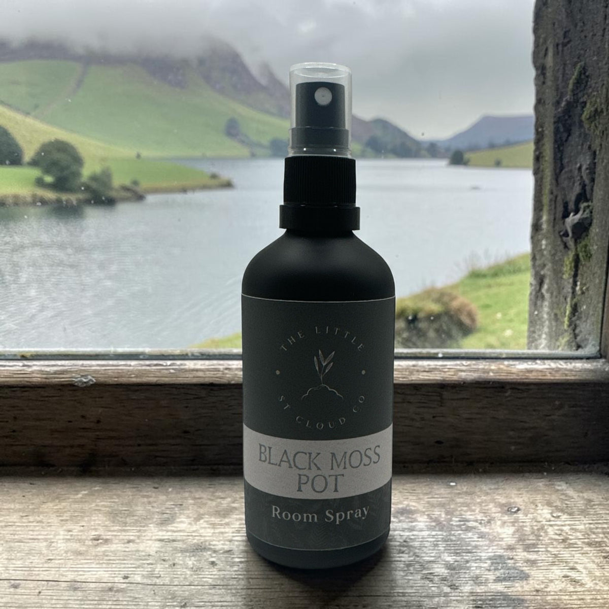 A matte black room spray bottle labeled 'The Little Cloud Co. Black Moss Pot' sitting on a rustic wooden windowsill with a blurred outdoor landscape of green hills and a lake in the background.