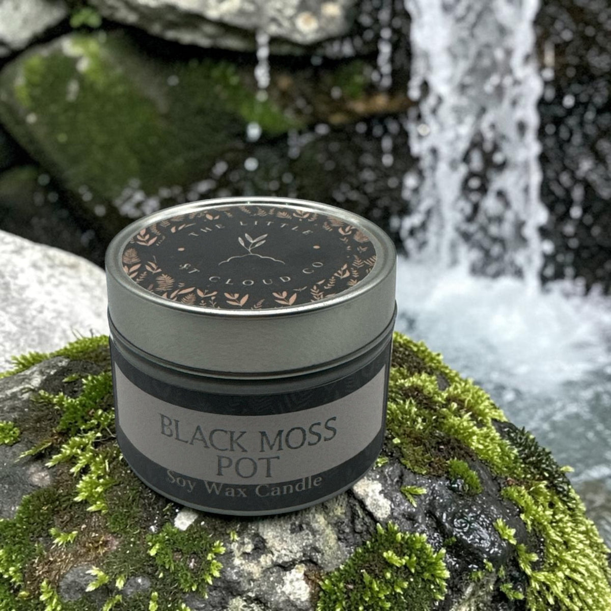 A jar candle labeled 'BLACK MOSS POT' sits on a mossy rock with a blurred waterfall and forest background. The candle jar is dark gray with a silver lid and a gray-and-black label.