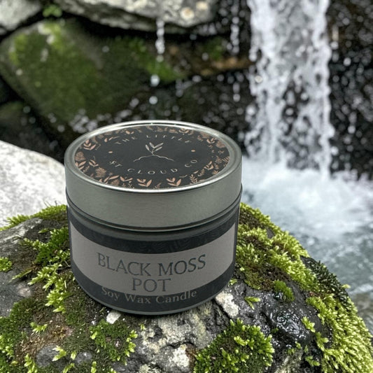 A jar candle labeled 'BLACK MOSS POT' sits on a mossy rock with a blurred waterfall and forest background. The candle jar is dark gray with a silver lid and a gray-and-black label.