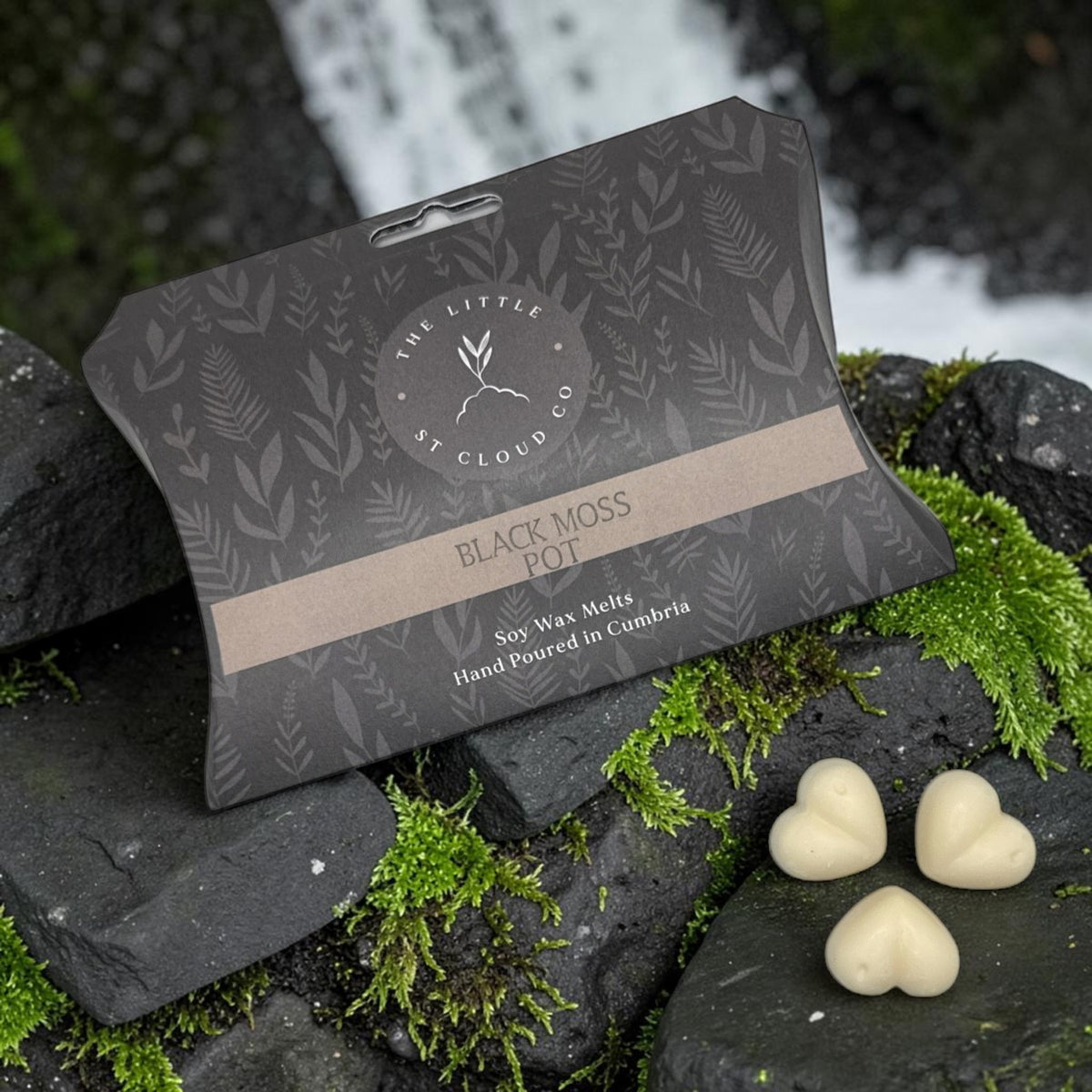 The Little St Cloud Co Black Moss Pot wax melts packaging displayed on mossy rocks next to three heart-shaped cream wax melts, with a blurred waterfall background.