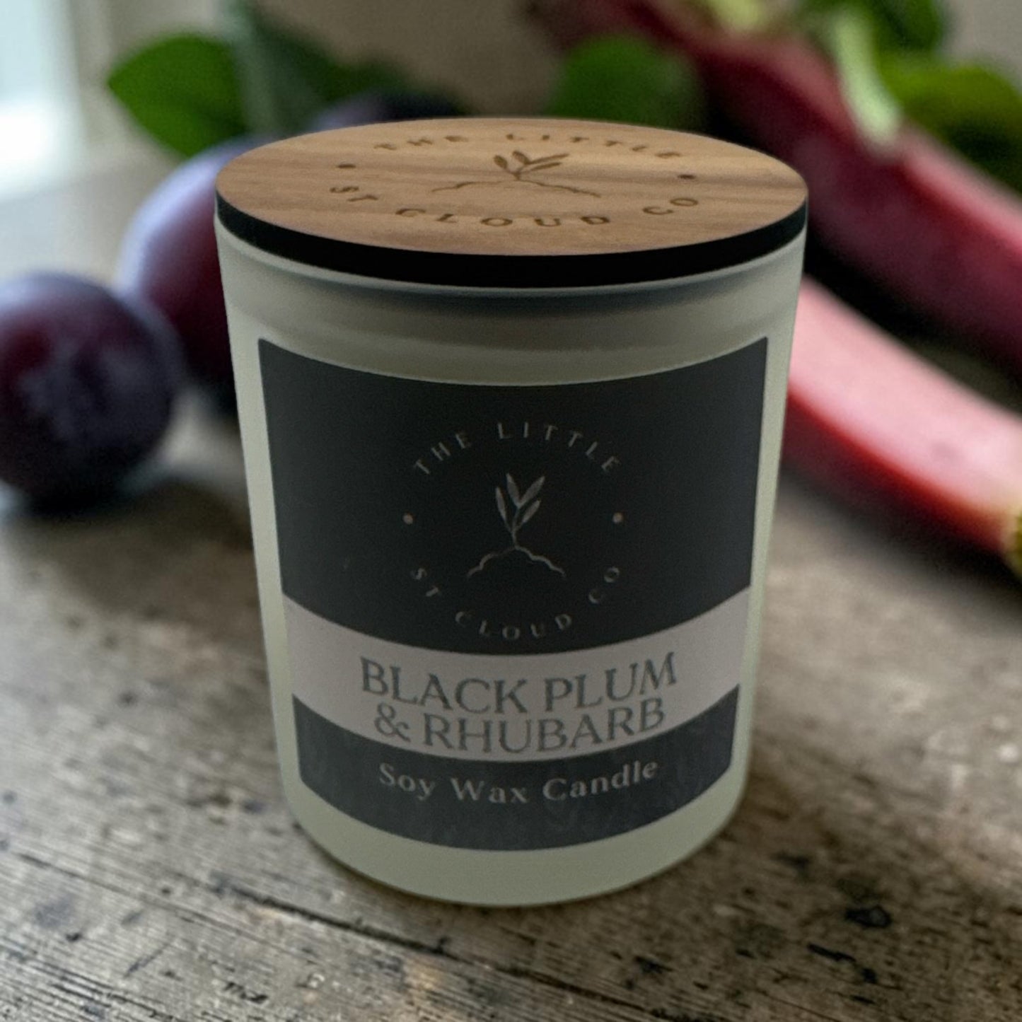 Soy wax candle labeled 'Black Plum & Rhubarb' with a wooden lid on a rustic surface.