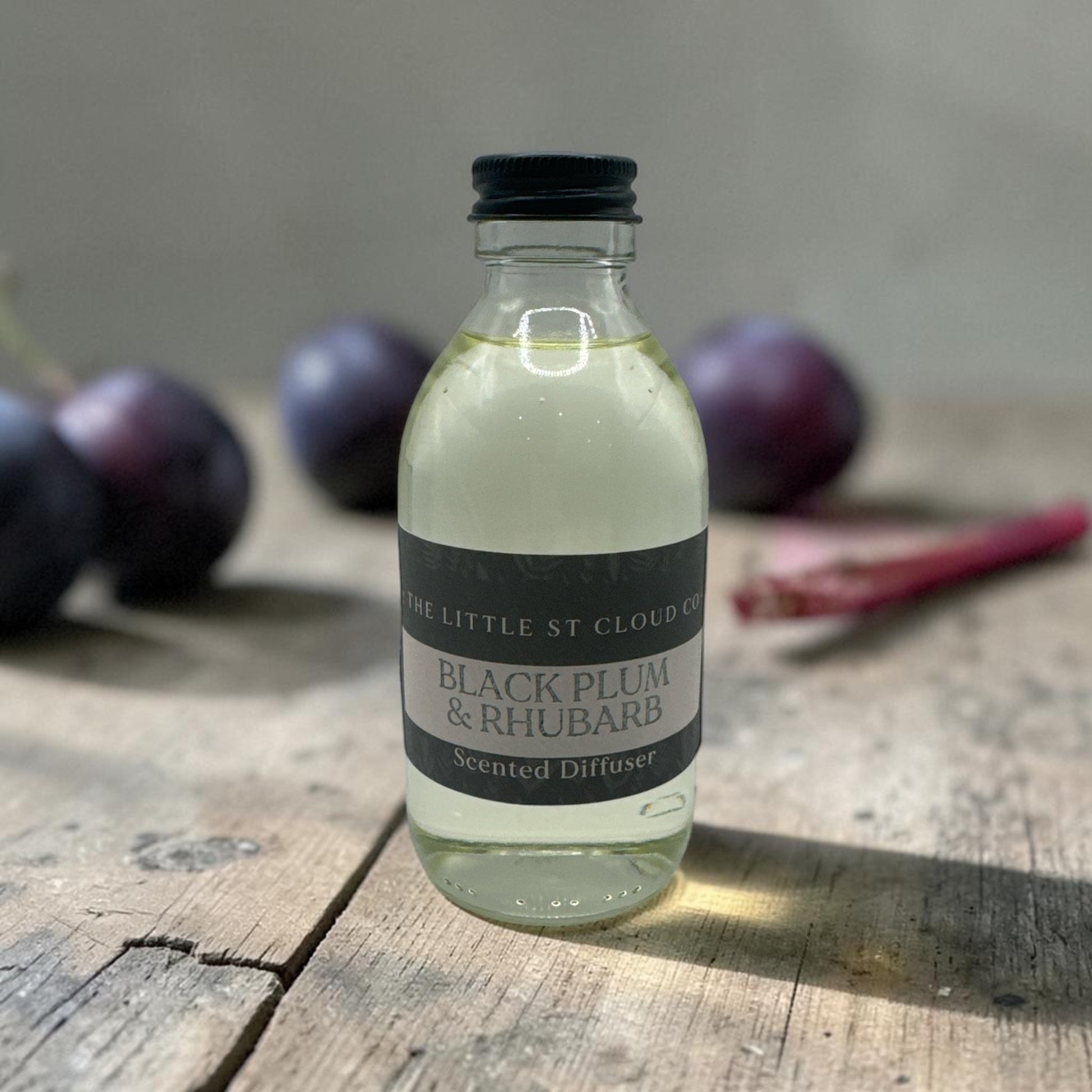 Bottle of 'Black Plum & Rhubarb' scented diffuser on a wooden surface with plums in the background.