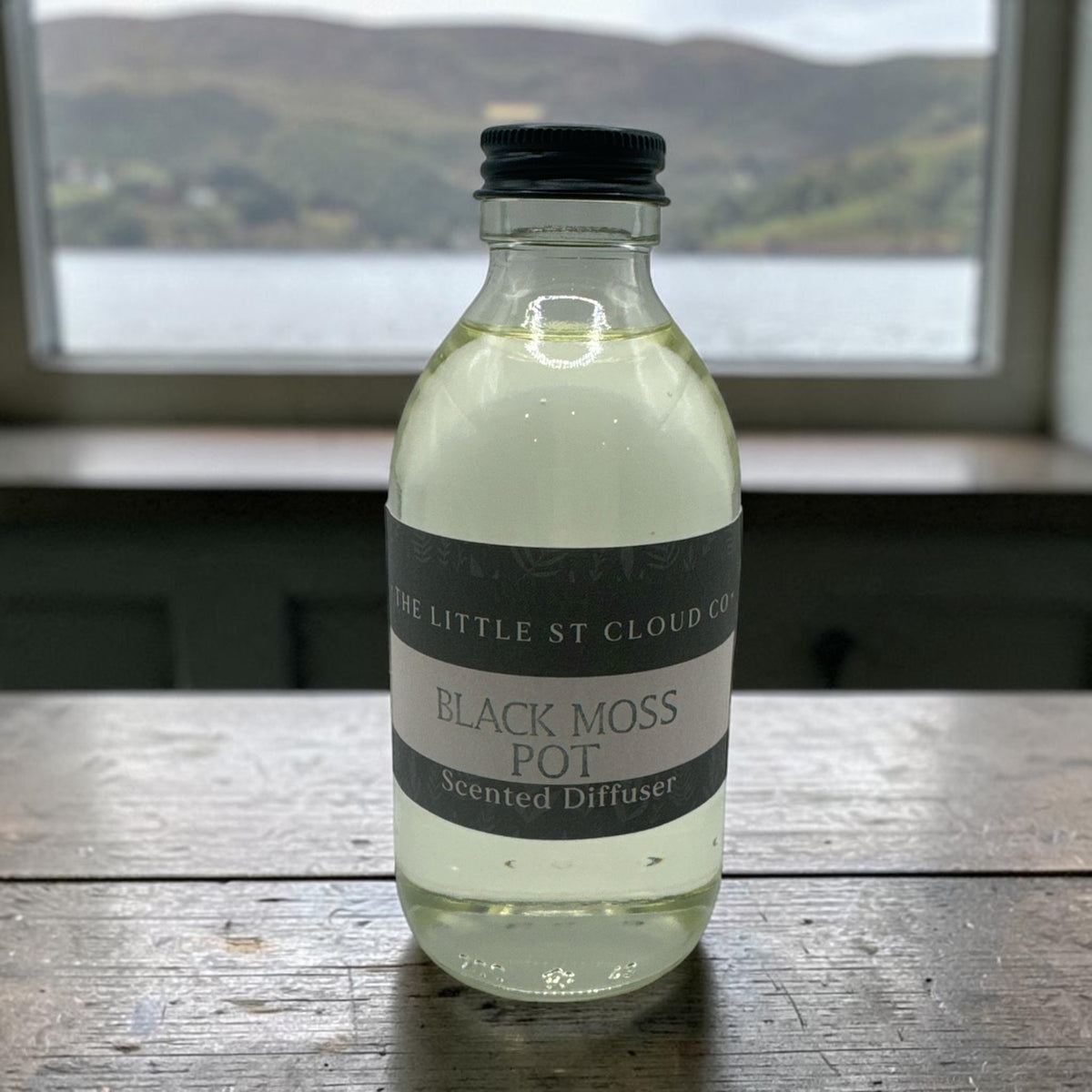 A clear glass bottle of 'Black Moss Pot' scented diffuser liquid by 'The Little St Cloud Co' with a black cap, sitting on a rustic wooden table in front of a window showing a blurred outdoor landscape.