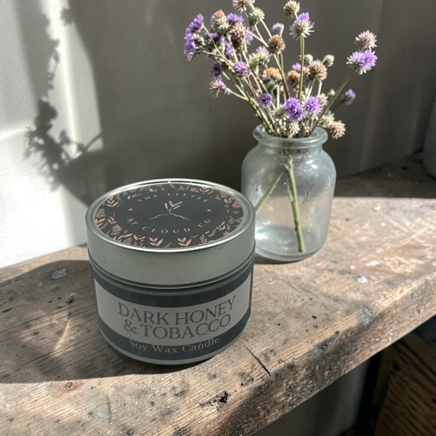 Candle labeled 'Dark Honey & Tobacco' on a wooden surface with a jar of flowers in the background.