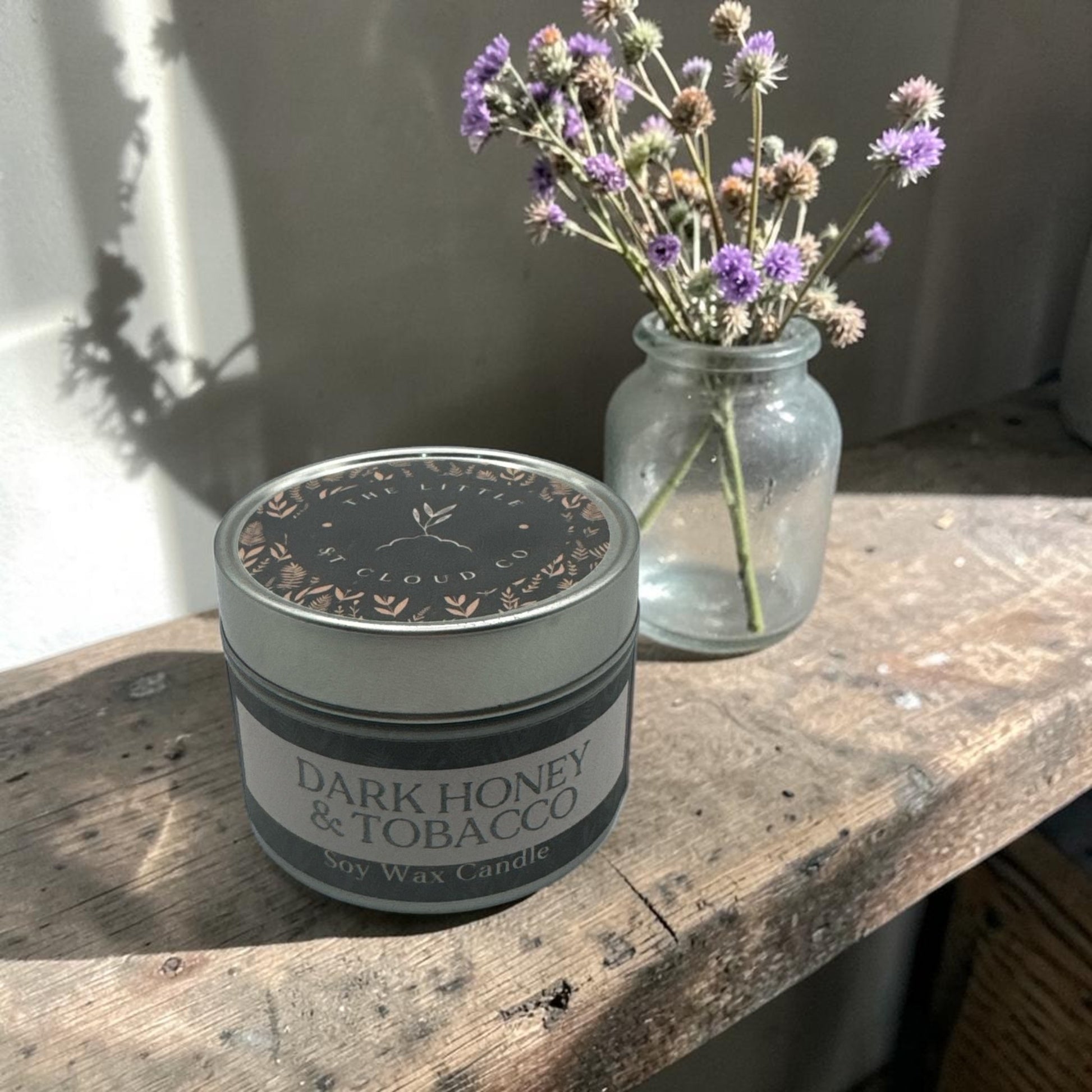 Candle labeled 'Dark Honey & Tobacco' on a wooden surface with a jar of flowers in the background.