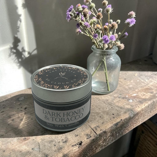 Candle labeled 'Dark Honey & Tobacco' on a wooden surface with a jar of flowers in the background.