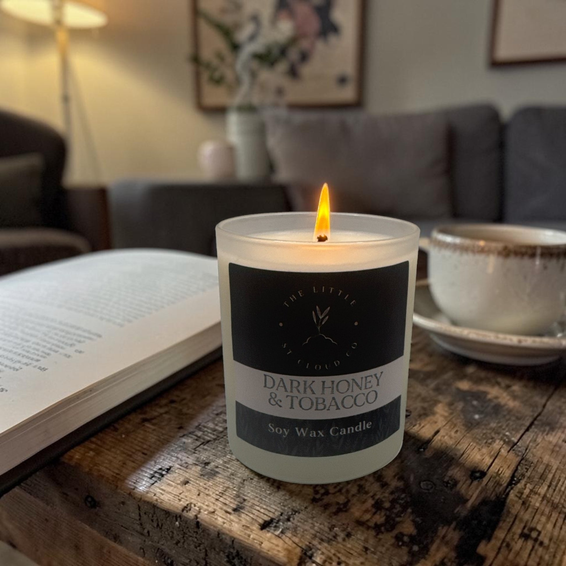 Candle labeled 'Dark Honey & Tobacco' on a wooden table with a book and cup in the background.