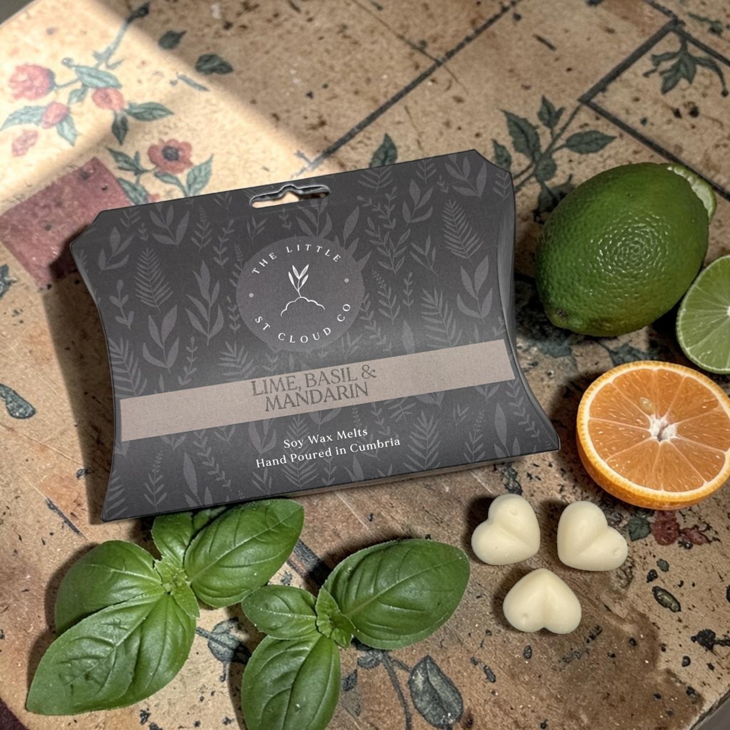 Packaged lime, basil, and mandarin wax melts on a decorative surface with fresh basil leaves and an orange.