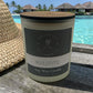 Soy wax candle labeled 'Maldives' with a straw hat on a wooden surface, tropical background.