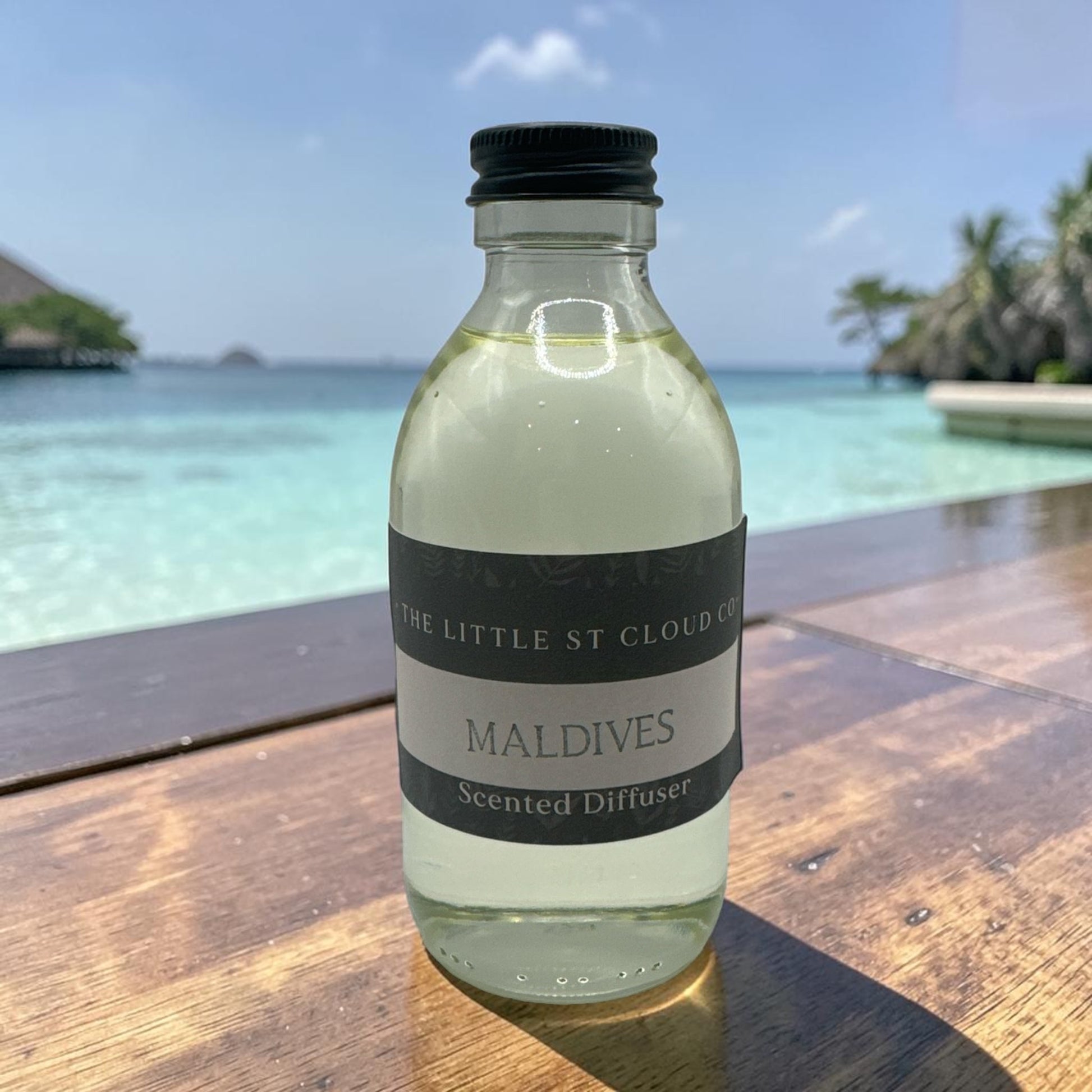 Bottle of 'The Little St Cloud Co' Maldives scented diffuser on a wooden table with a tropical background.