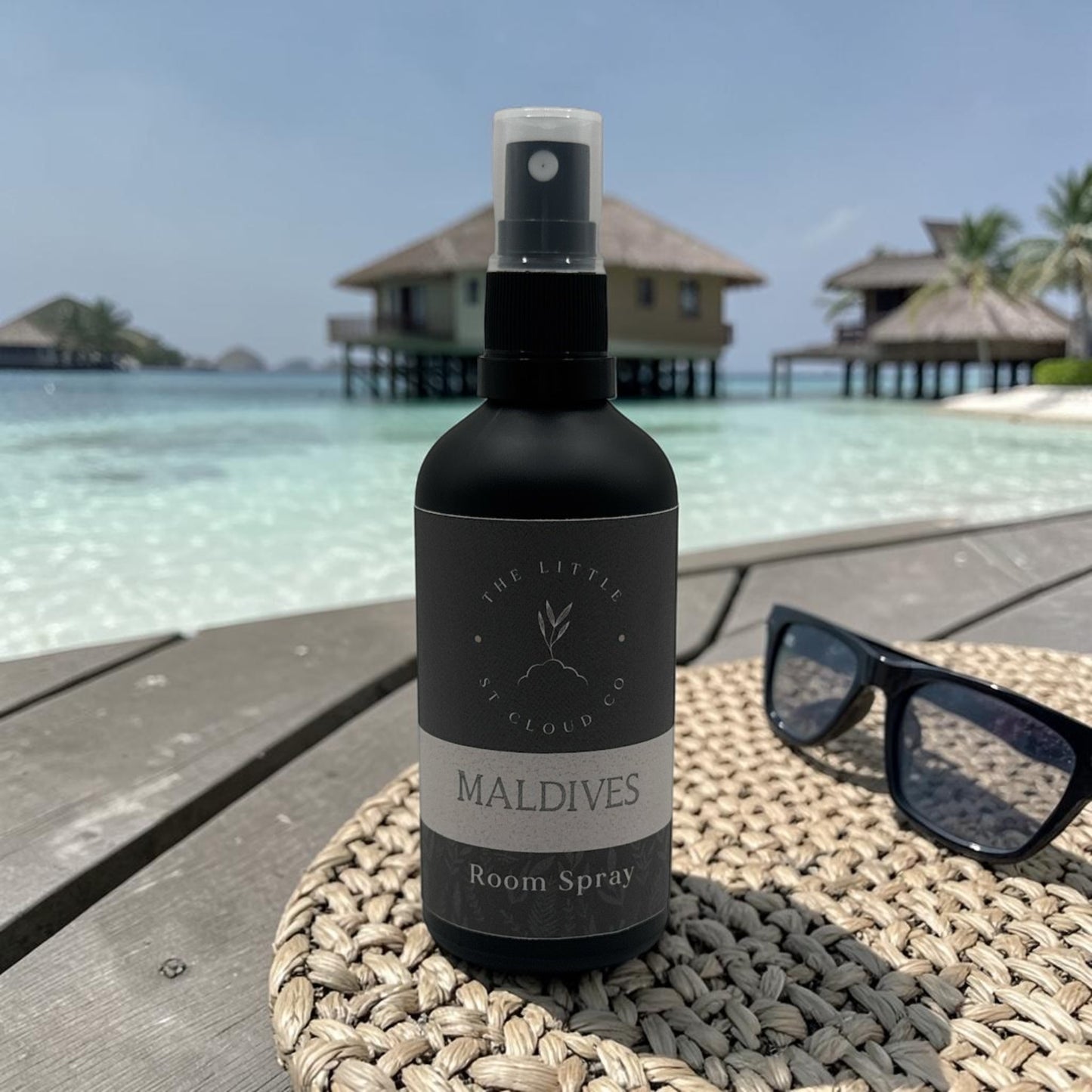 Room spray bottle labeled 'Maldives' on a woven mat with a tropical resort background