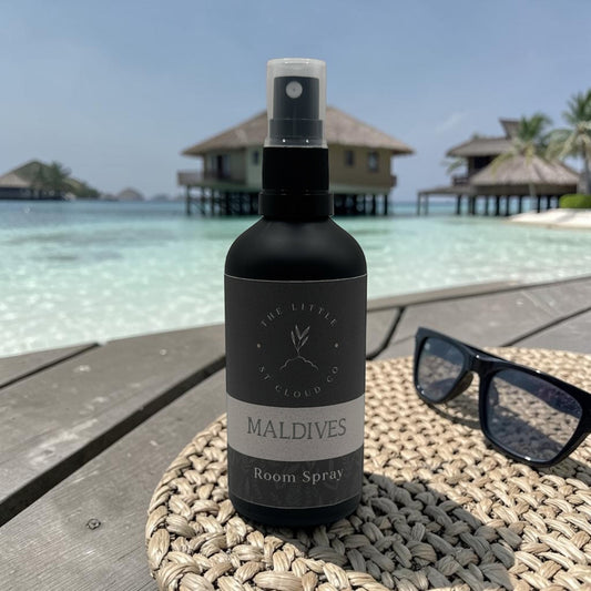 Room spray bottle labeled 'Maldives' on a woven mat with a tropical resort background