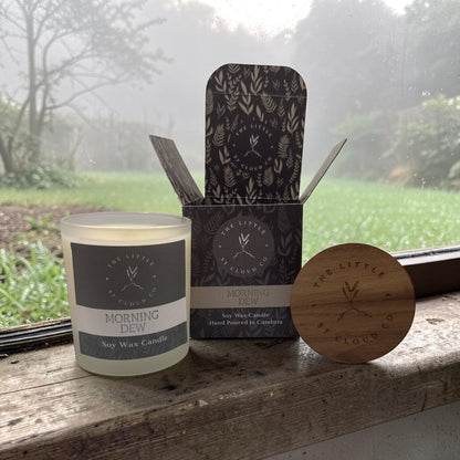 Soy wax candle with packaging and wooden lid on a windowsill with a misty garden view.