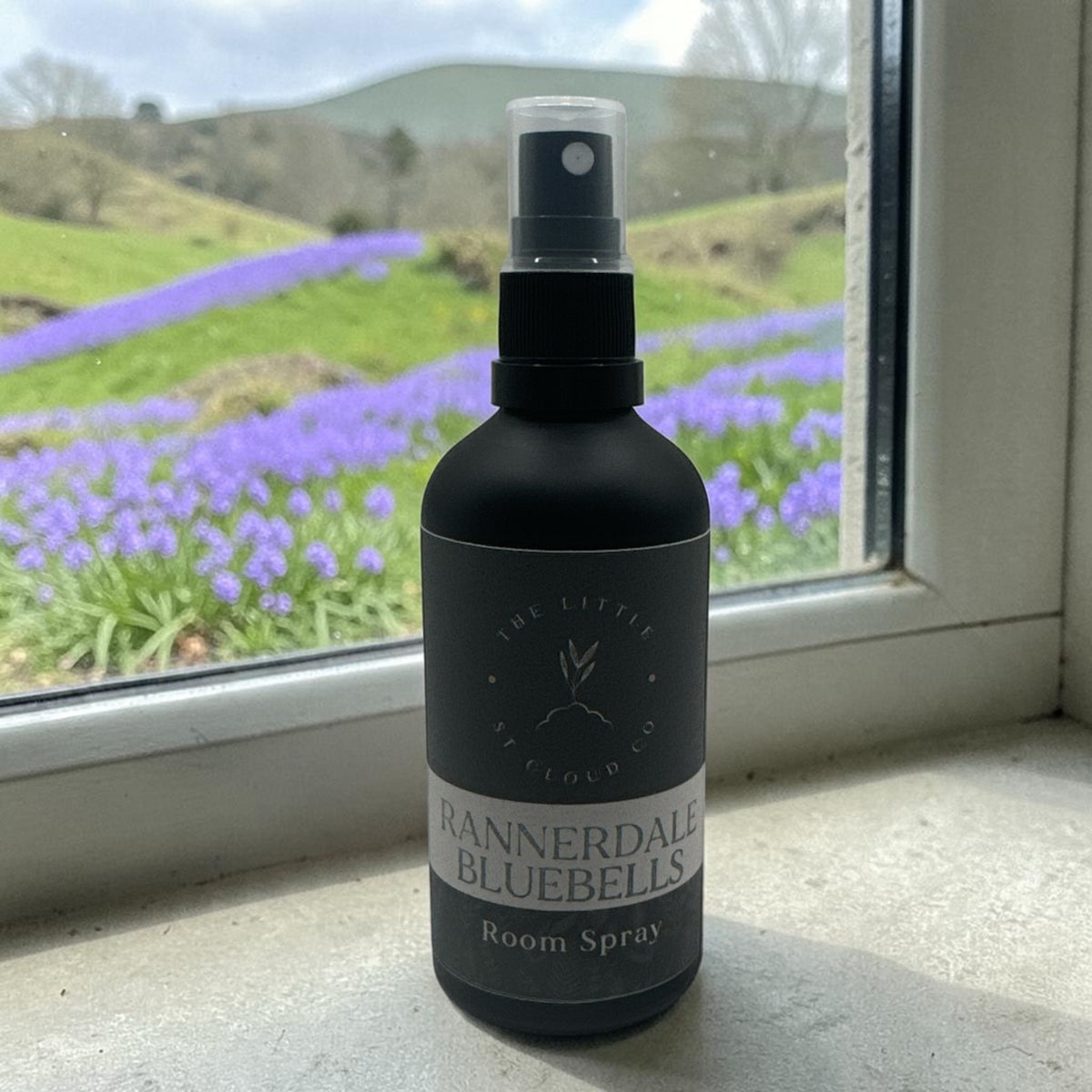 A Matte black room spray in the scent Rannerdale Bluebells by the little st cloud company in a windowsil with views of bluebells