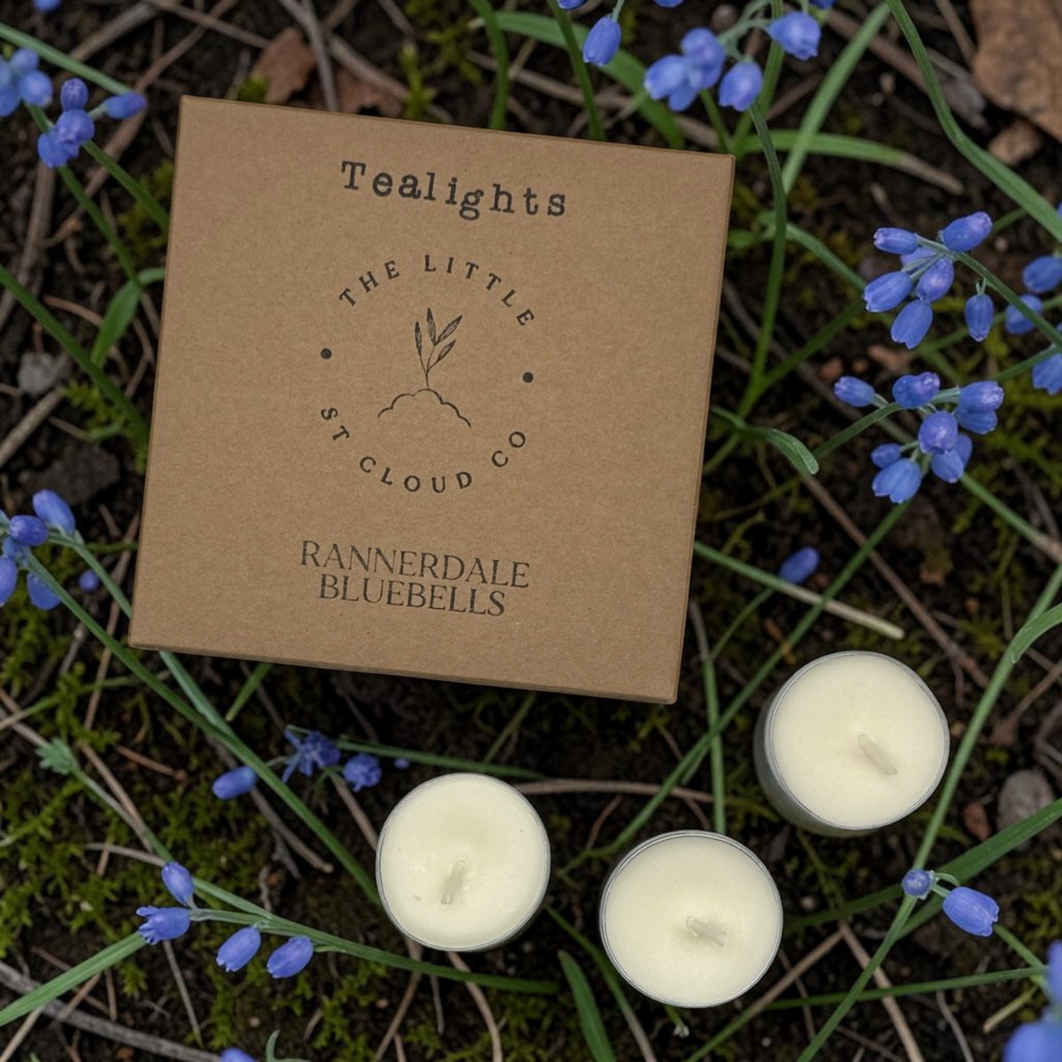 A square kraft paper box labeled 'Tealights' and 'Rannerdale Bluebells' by The Little St Cloud Co, placed outdoors among bluebells and green grass with three tealight candles visible.