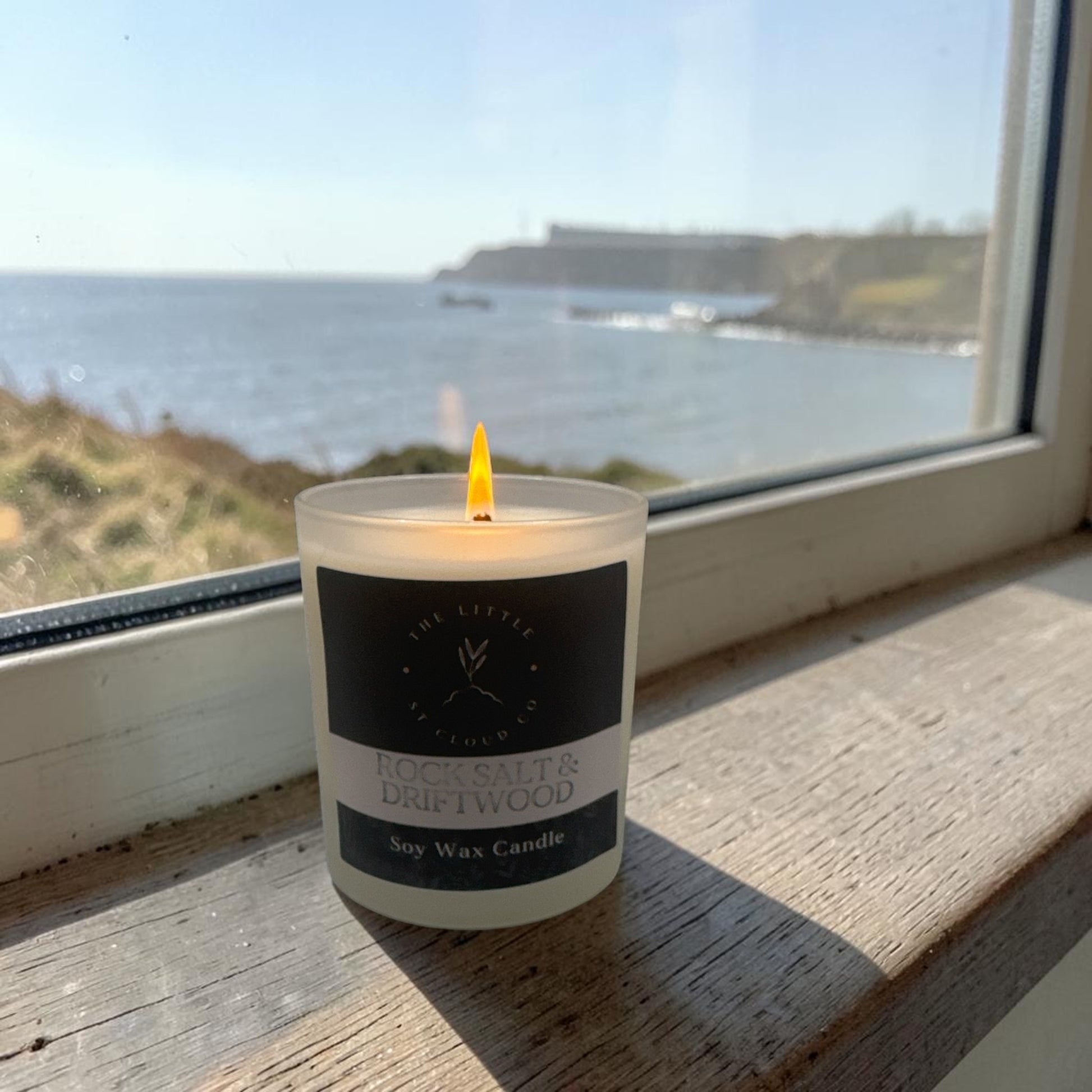 Candle labeled 'Rock Salt & Driftwood' on a windowsill with a scenic view of water and mountains.
