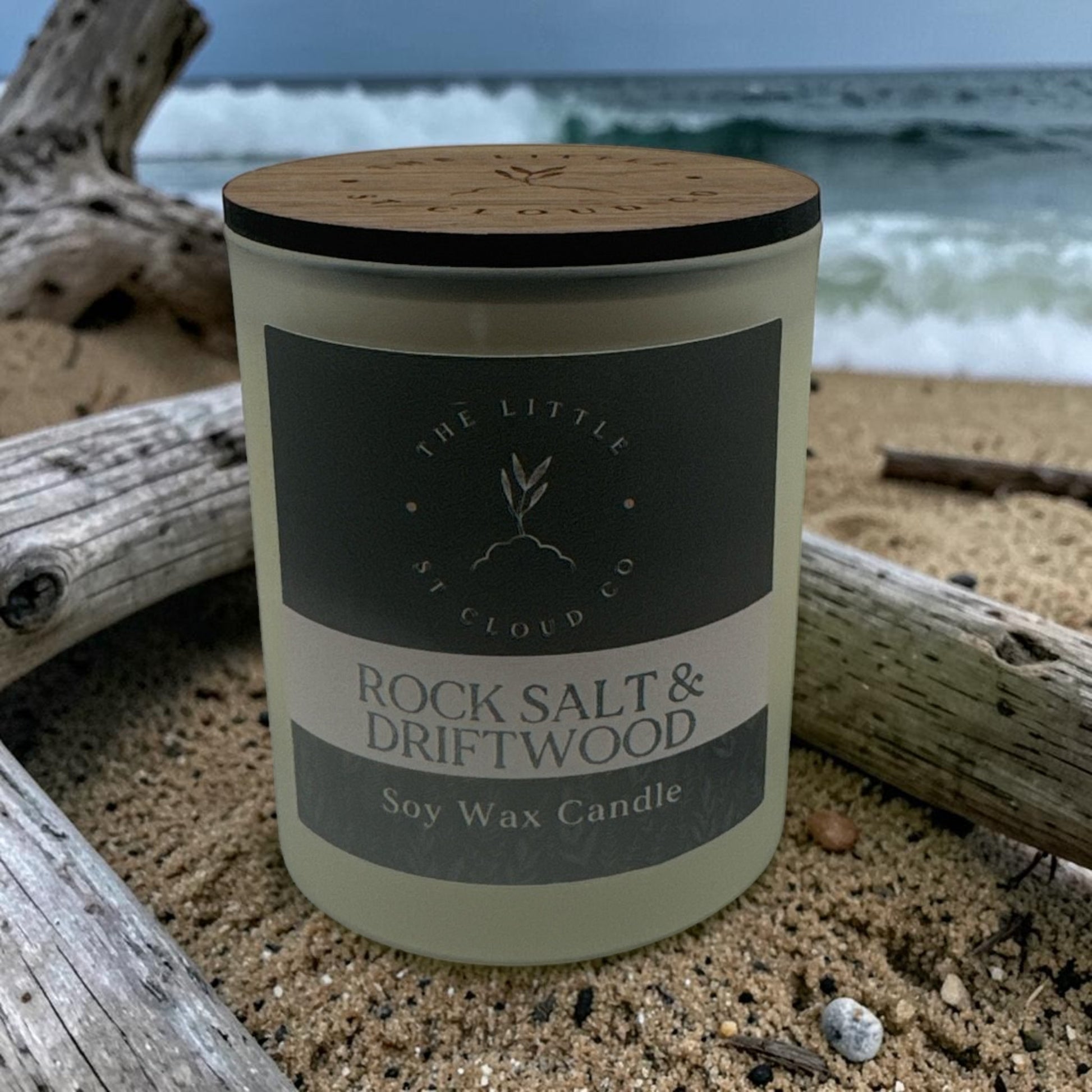 Candle labeled 'Rock Salt & Driftwood' on a beach with ocean waves in the background