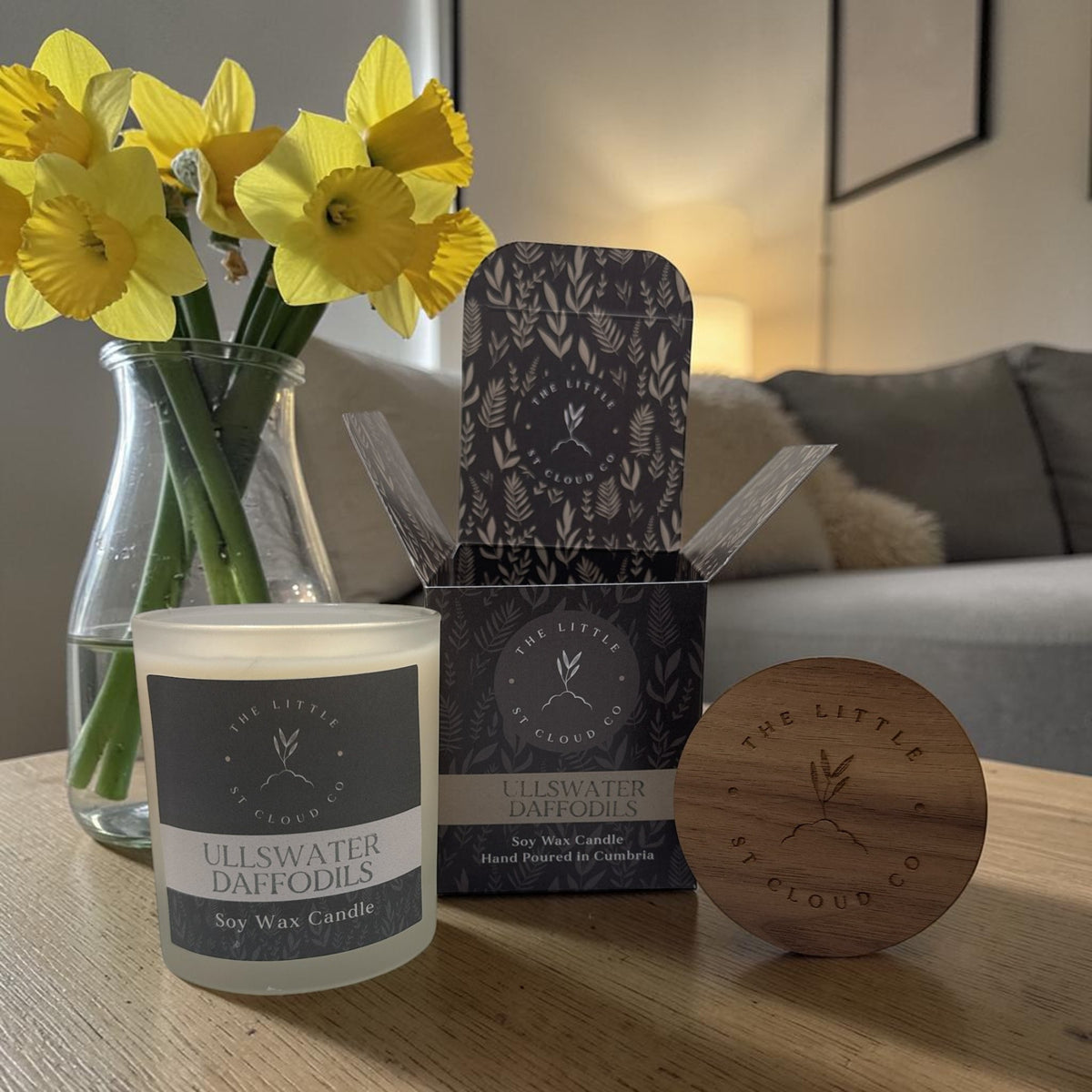 Frosted candle jar in the scent ullswater daffodils next to a gift box and wooden lid in a cosy living room next to a vase of daffodils