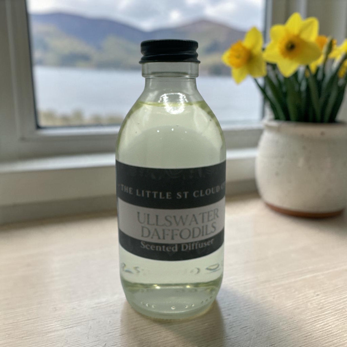 A clear glass bottle of diffuser refill in the scent ullswater daffodils with a black lid in front of a lakeside view with a vase of daffodils