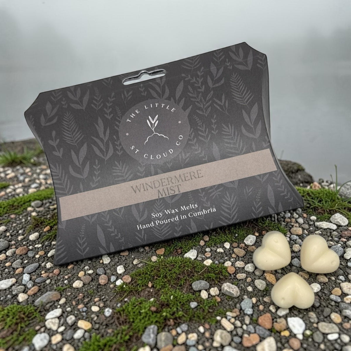 A grey branded box of wax melts and three heart shaped wax melts by the little st cloud co in the scent windermere mist an o gravel bed with mist lake views